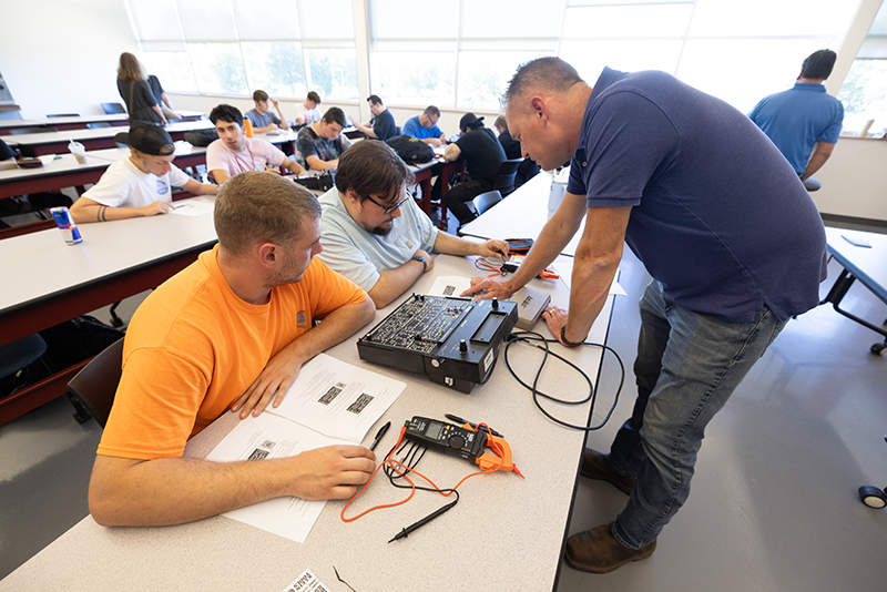 An instructor leans over a table to show two students how a piece of industrial maintenance technology works