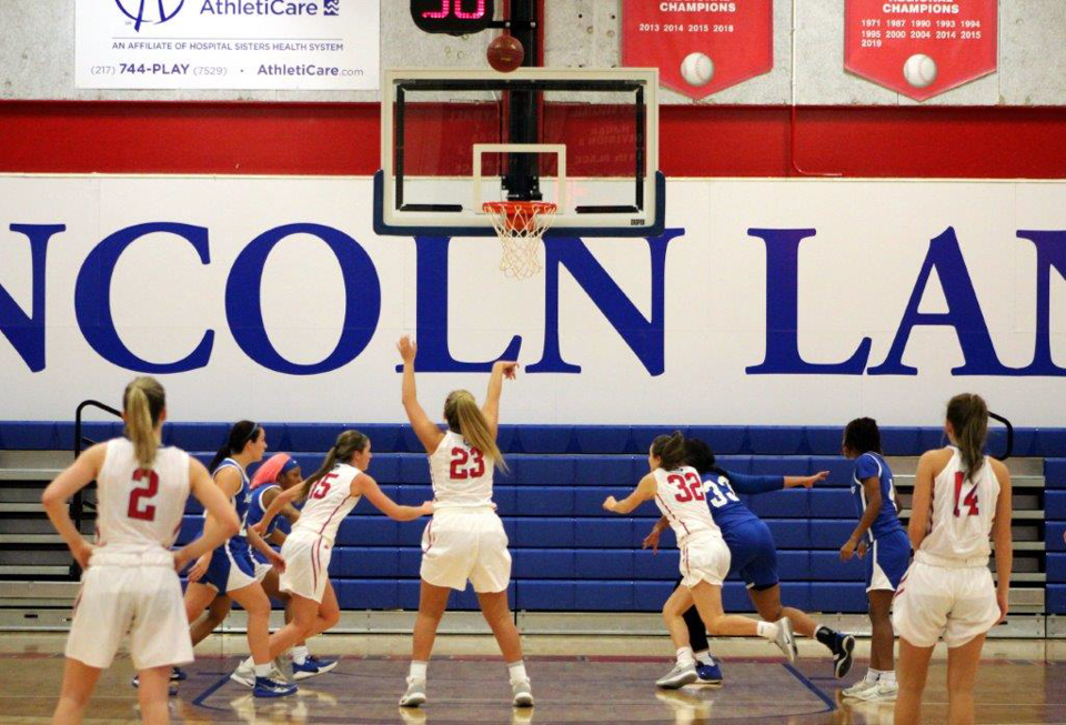 Several LLCC women's basketball players gather around the basket after a teammate has shot the ball toward the net. 