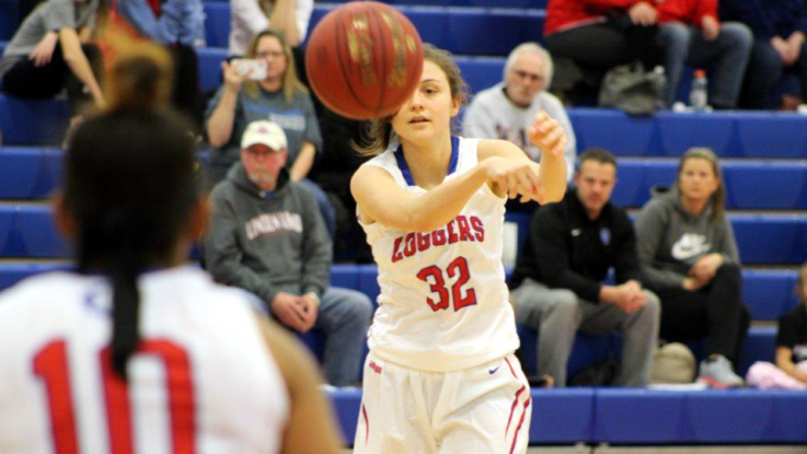 An LLCC women's basketball player in a white uniform throws a ball towards a fellow player while a crowd sits behind her in the bleachers. 