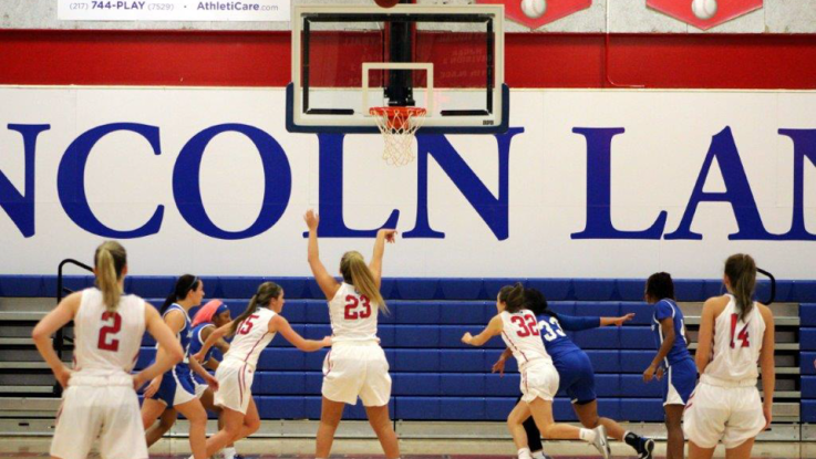 Several LLCC women's basketball players gather around the basket after a teammate has shot the ball toward the net. 