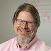 A man with gray-ish hair and glasses, wearing a light-pink colored shirt.