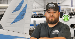 Man in an LLCC aviation black hat and tshirt standing near the tail of an airplane.